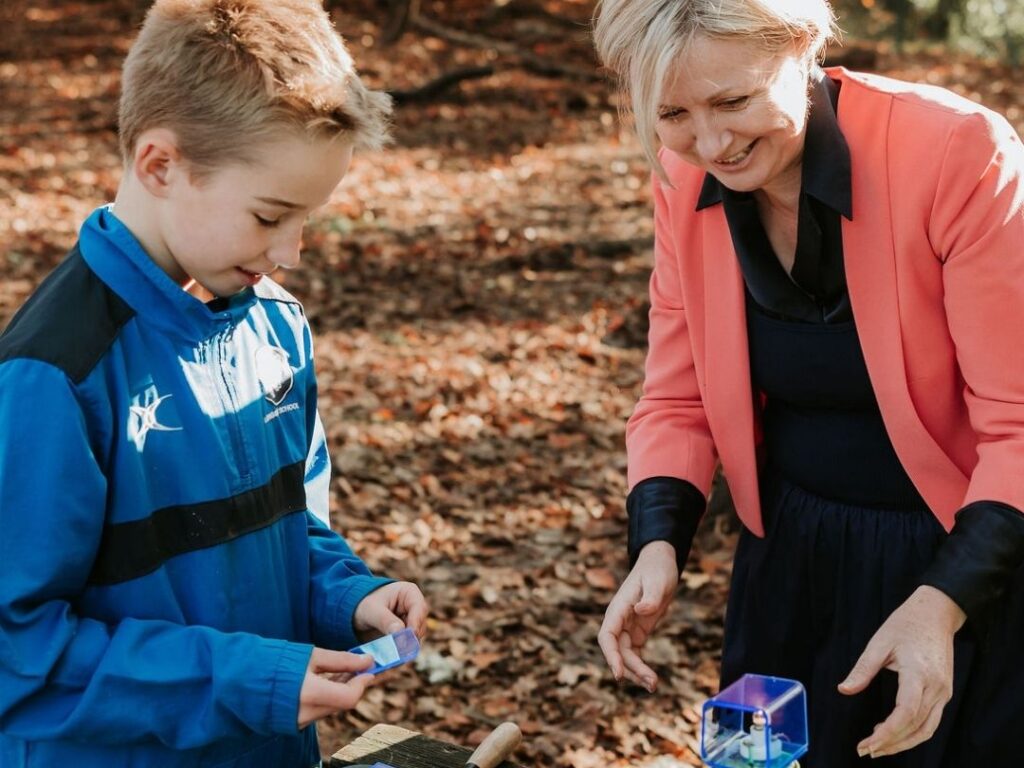 Head teacher outdoors in the woods with a student, looking at and discussing his DT work, both engaged and smiling.
