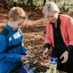 Head teacher outdoors in the woods with a student, looking at and discussing his DT work, both engaged and smiling.